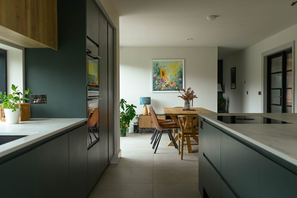 A modern kitchen with deep green cabinetry, light countertops, and an integrated oven, leading into a bright dining space with a wooden table, tan leather chairs, and colourful artwork on the wall. Potted plants and natural textures add warmth to the design.