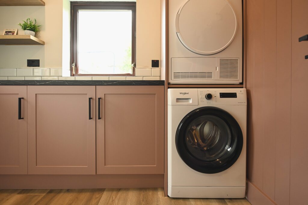 A modern pink utility room featuring stacked Whirlpool washer and dryer, shaker-style pink cabinetry with black handles, a dark worktop, and a natural wood floor.
