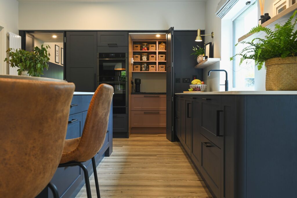 A hidden pantry revealed inside a navy kitchen, featuring soft pink cabinetry, natural wood shelves, and woven storage baskets for a warm and modern contrast.