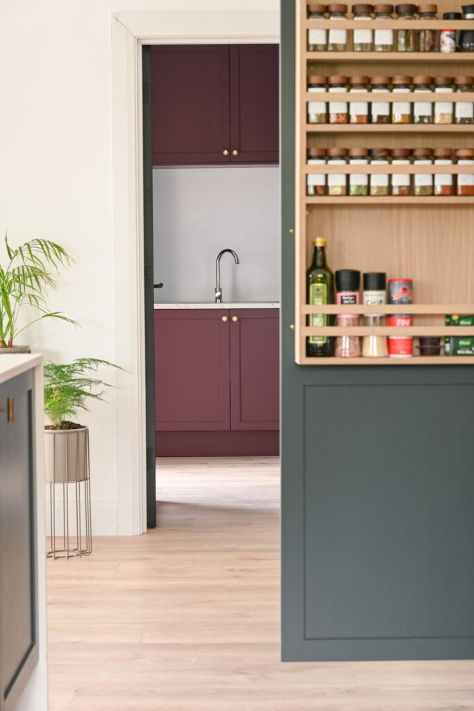 Bespoke shaker kitchen with oak spice storage, deep forest green cabinetry and a view into the adjoining plum Shaker utility room.