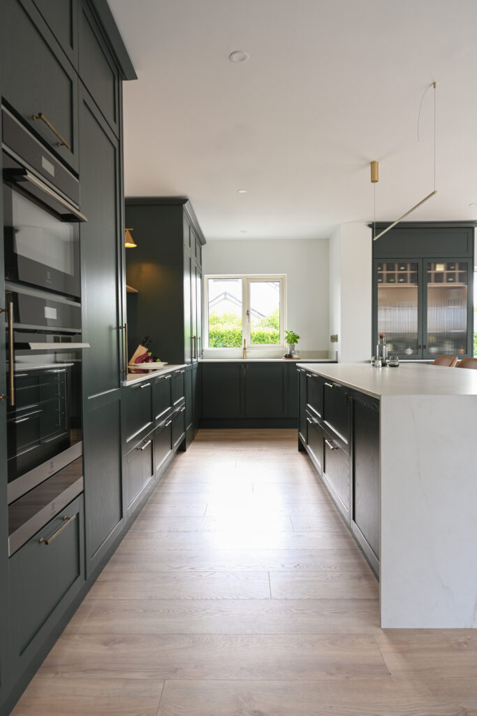 Deep forest green Shaker kitchen extension with integrated ovens, Dekton Rem waterfall island, brass hardware and bespoke reeded-glass bar cabinetry.