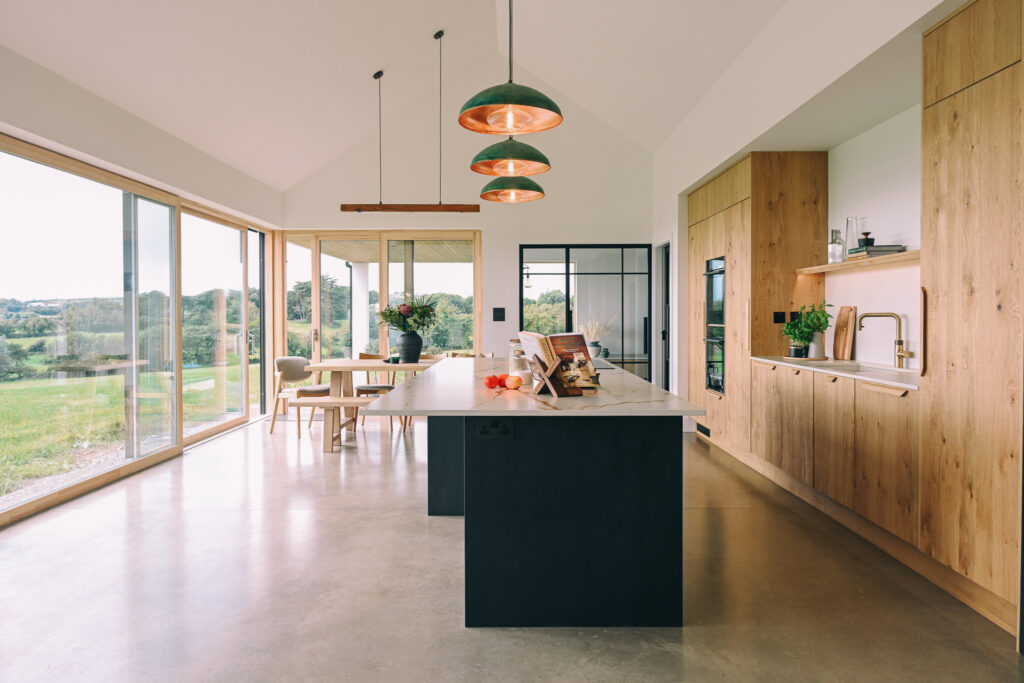 Bespoke oak veneer kitchen with contrasting black island, Dekton Reverie worktop, Quooker tap and floor-to-ceiling glazing framing the coastal views.