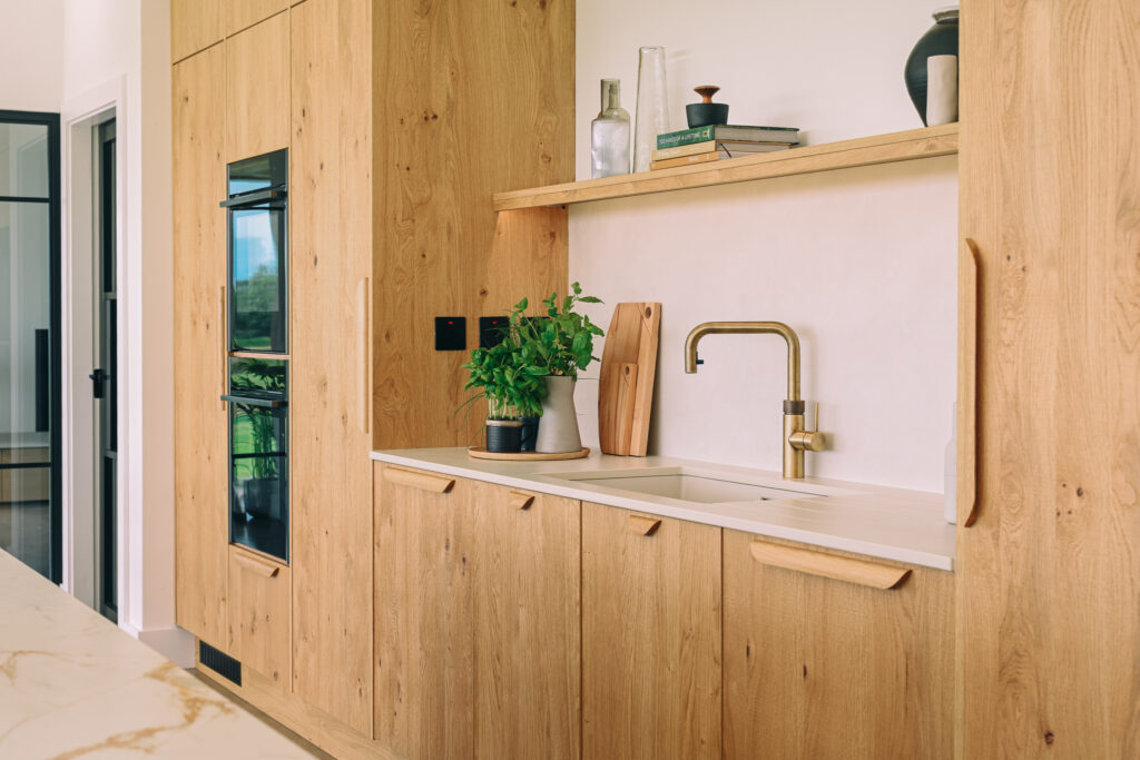Bespoke oak kitchen cabinetry with integrated ovens, Quooker tap, open shelf and light stone worktop.